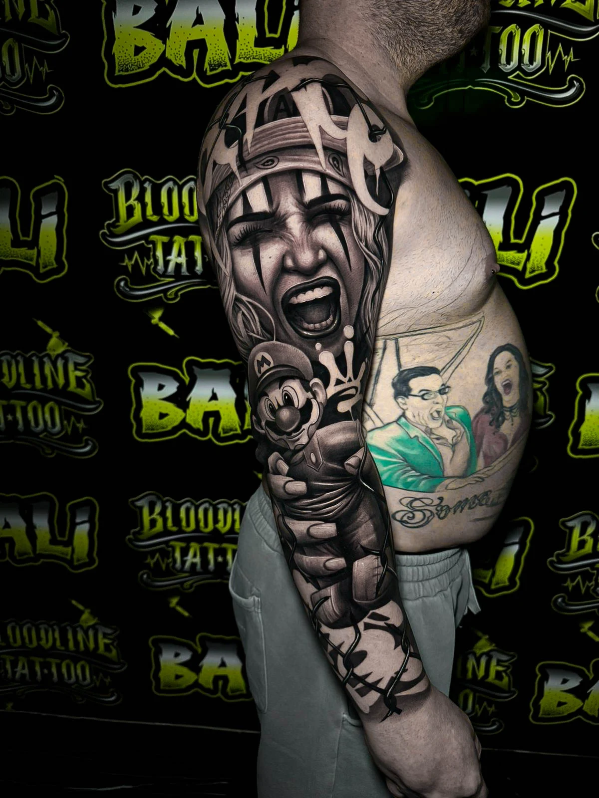 Detailed black and grey Chicano upper arm sleeve tattoo featuring a screaming clown girl, Mario figure, and bold graffiti-style script at Bloodline Tattoo Bali.