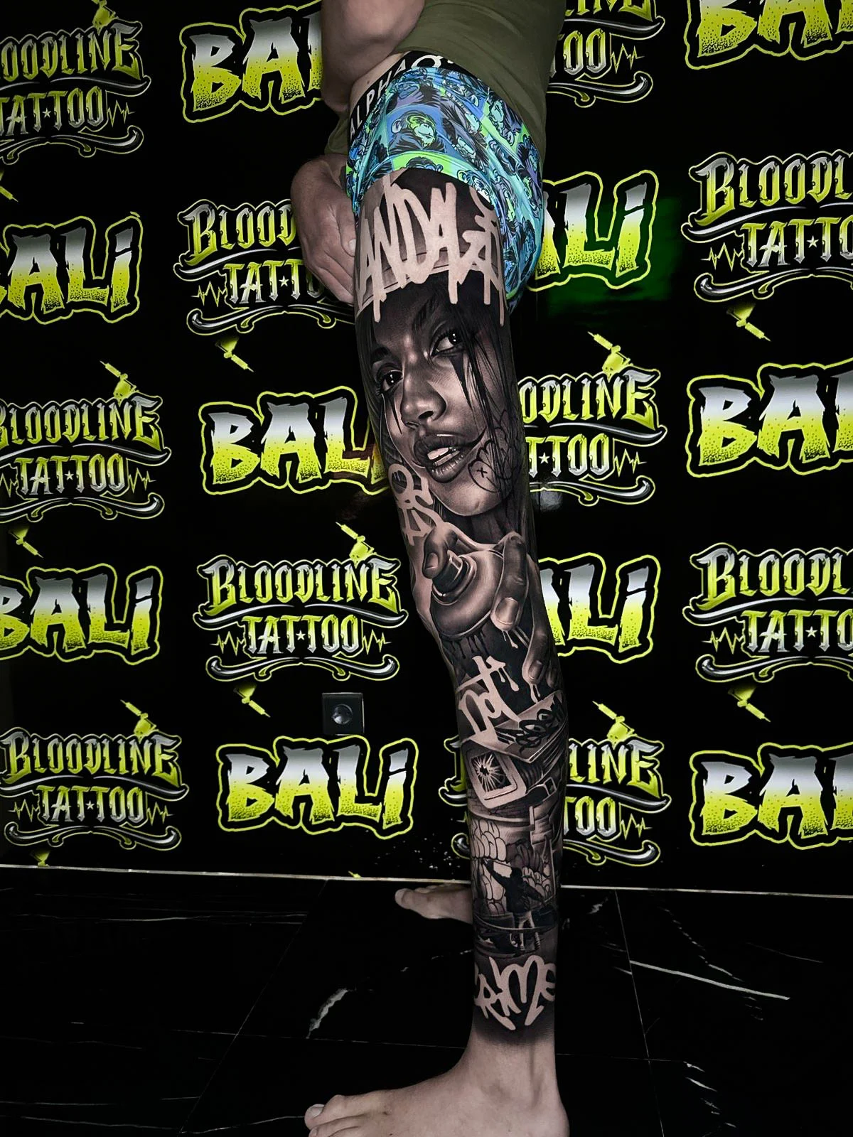 Detailed black and grey Chicano leg sleeve tattoo featuring a female portrait, spray can, block lettering, and urban street imagery at Bloodline Tattoo Bali.