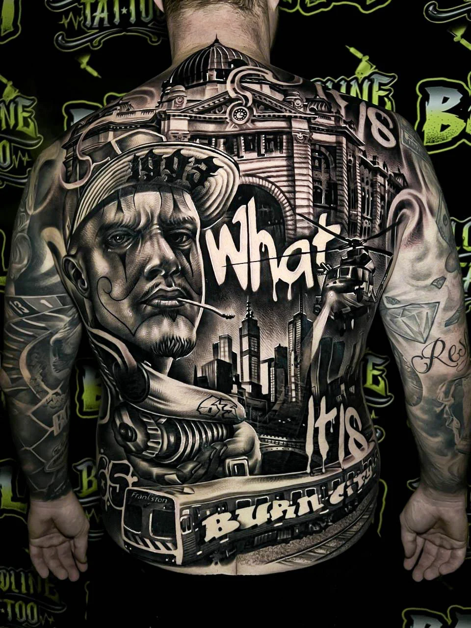 Detailed black and grey Chicano full back tattoo with a gangster portrait, city skyline, helicopter, tram, and large script lettering at Bloodline Tattoo Bali.