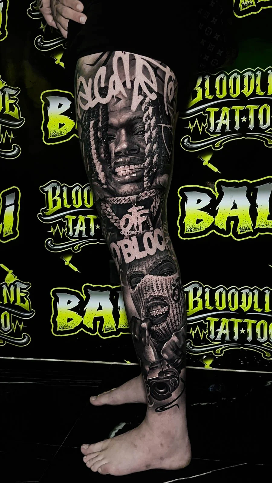 Detailed black and grey Chicano thigh and shin tattoo with a braided rapper portrait, bold script, ski mask figure, and handgun at Bloodline Tattoo Bali.