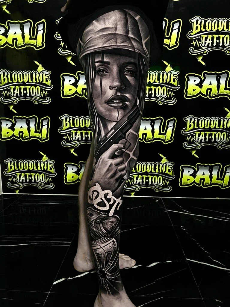 Detailed black and grey Chicano leg sleeve tattoo of a woman in a cap holding a pistol with cash and a nun portrait below at Bloodline Tattoo Bali.