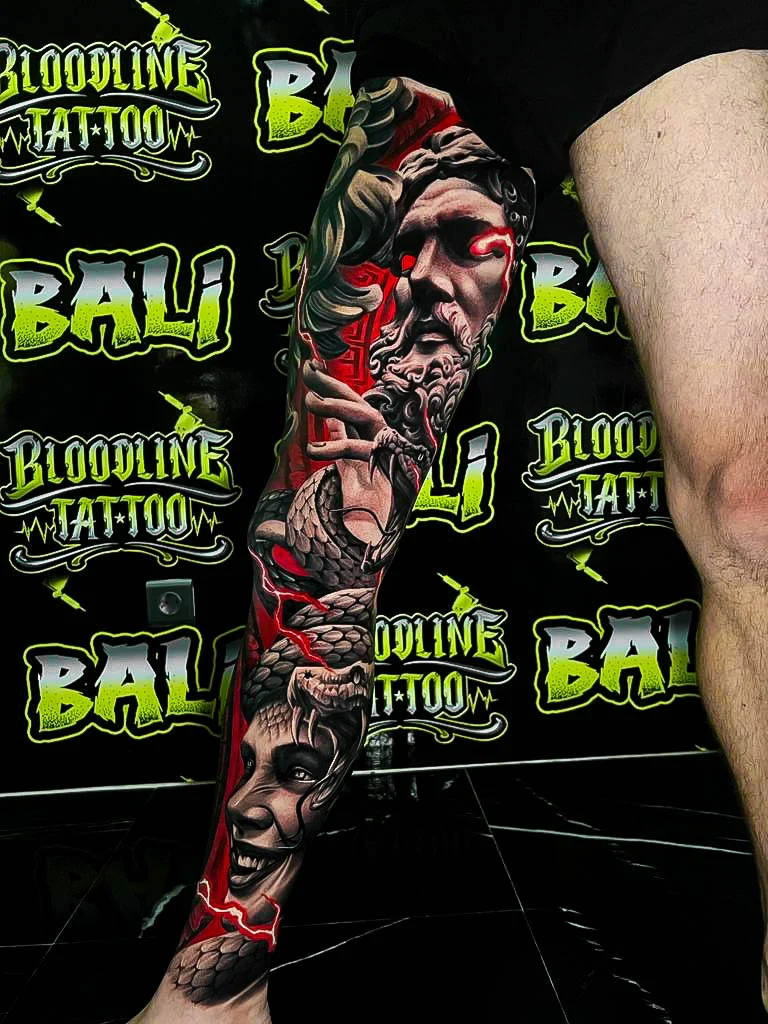 Colour leg sleeve tattoo featuring a red-eyed classical statue, coiling snake scales, and a sinister grinning face at Bloodline Tattoo Bali.