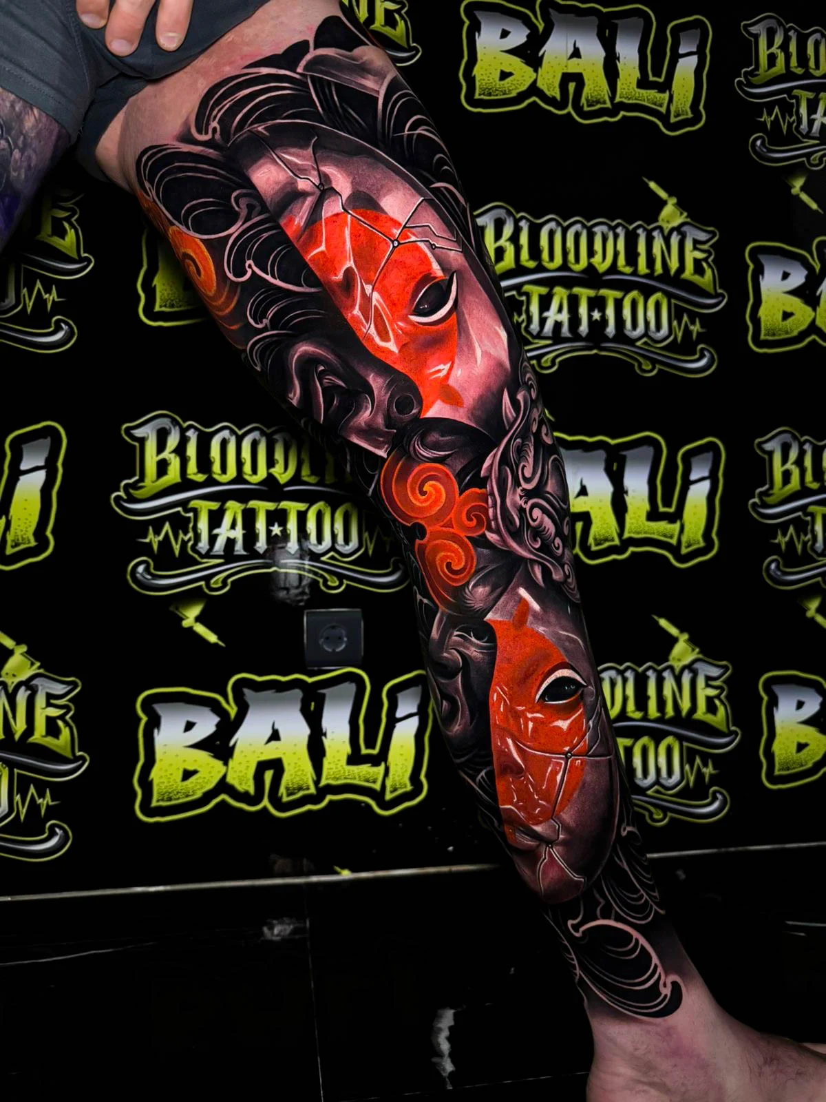 Colour thigh tattoo with cracked red theatrical masks, flowing black hair forms, and glowing orange cloud accents at Bloodline Tattoo Bali.