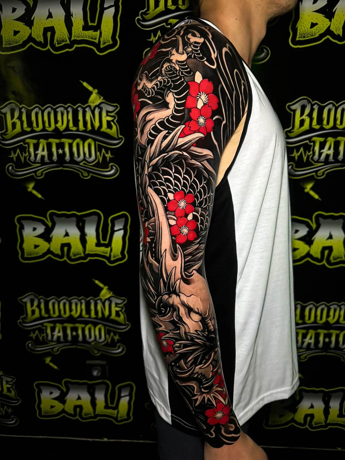 Black and grey Japanese full sleeve with dragon scales, skull details, flowing wind bars, and red cherry blossoms at Bloodline Tattoo Bali.