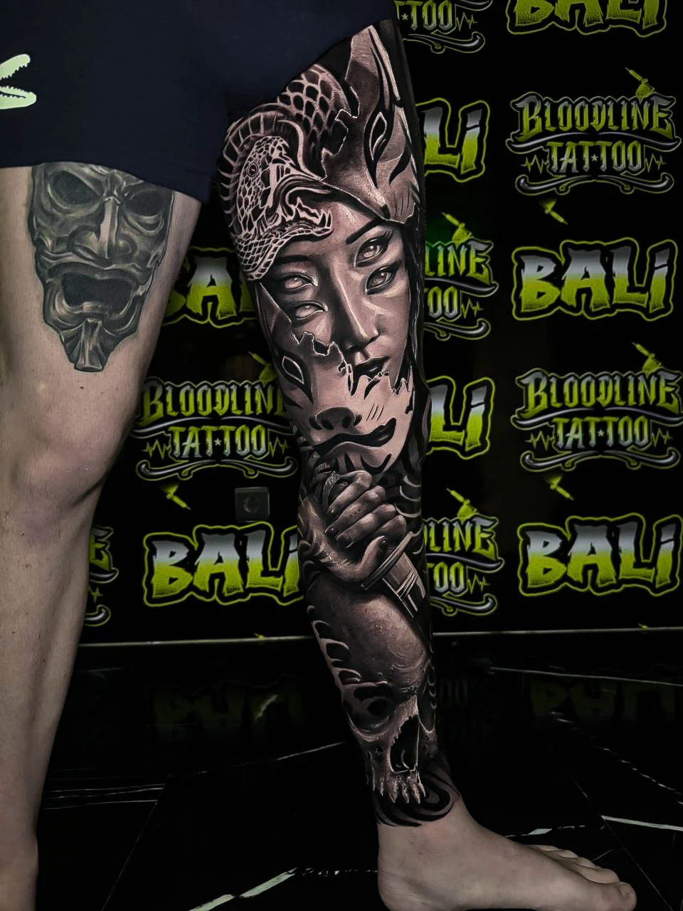 Black and grey Japanese lower leg tattoo with a kitsune mask, female portrait, clasped hands, and skull shading at Bloodline Tattoo Bali.