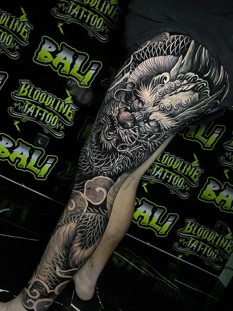 Black and grey Japanese full leg sleeve with a coiled dragon, wind bars, and crashing wave motifs at Bloodline Tattoo Bali.