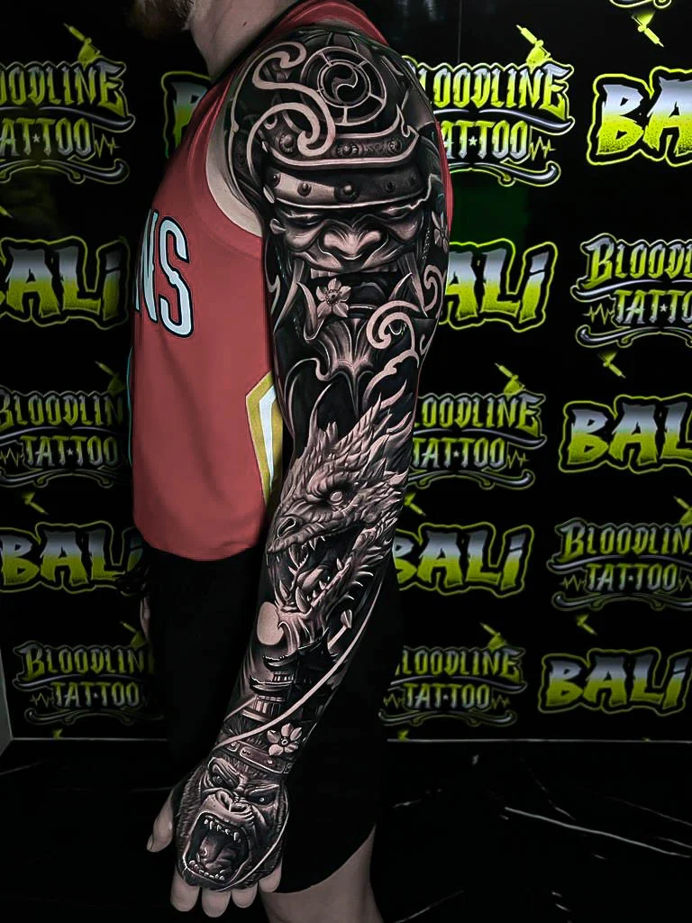 Black and grey Japanese full sleeve with a samurai kabuto, dragon head, cloud bars, and a snarling hand mask at Bloodline Tattoo Bali.