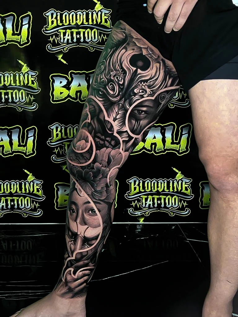 Black and grey Japanese leg sleeve with kitsune masks, peony blooms, and swirling wind bars at Bloodline Tattoo Bali.