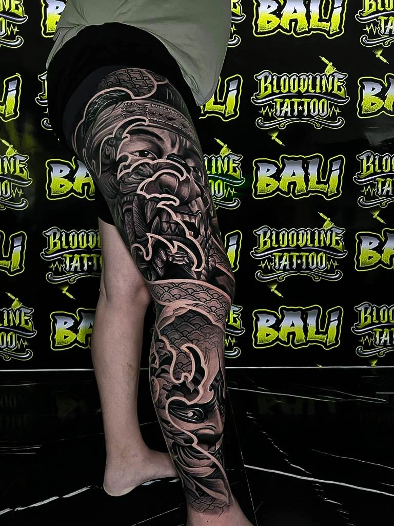 Black and grey Japanese full leg sleeve with a fierce warrior face, scale patterns, and wave bars at Bloodline Tattoo Bali.
