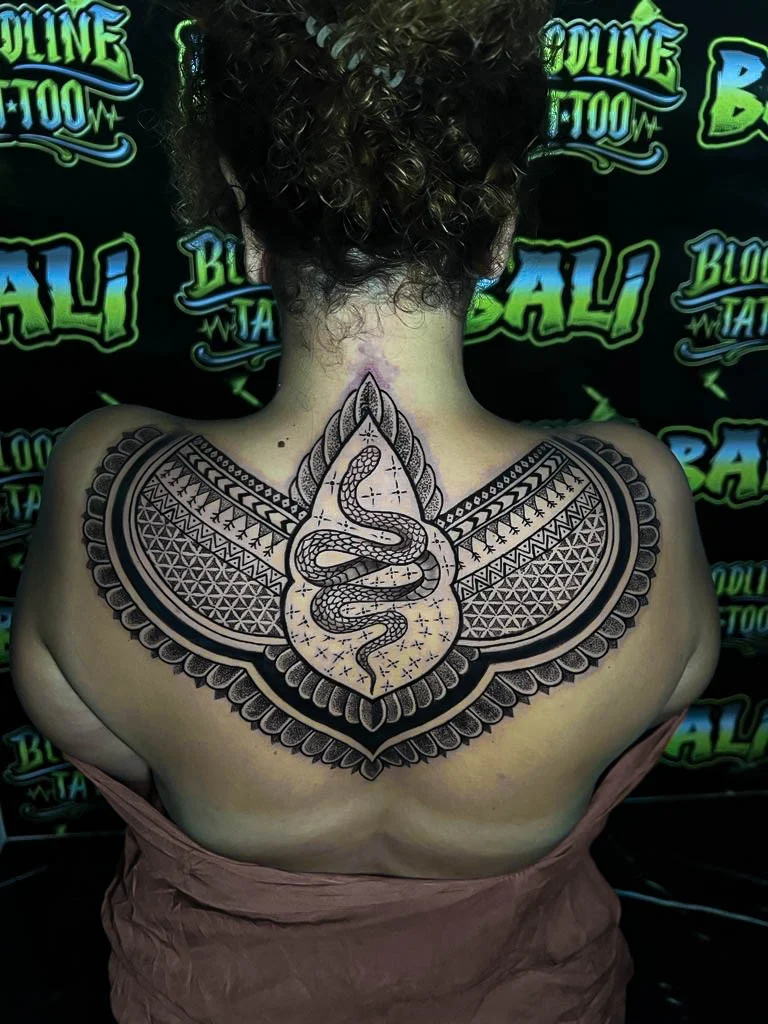 Upper-back blackwork with a coiled snake centerpiece, winged ornamental panels, and dense geometric patterning across both shoulders at Bloodline Tattoo Bali.