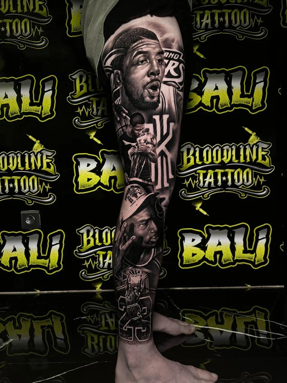 Black and grey portrait realism leg sleeve tattoo of a basketball player portrait with action scenes, jersey details, and championship-style imagery at Bloodline Tattoo Bali.