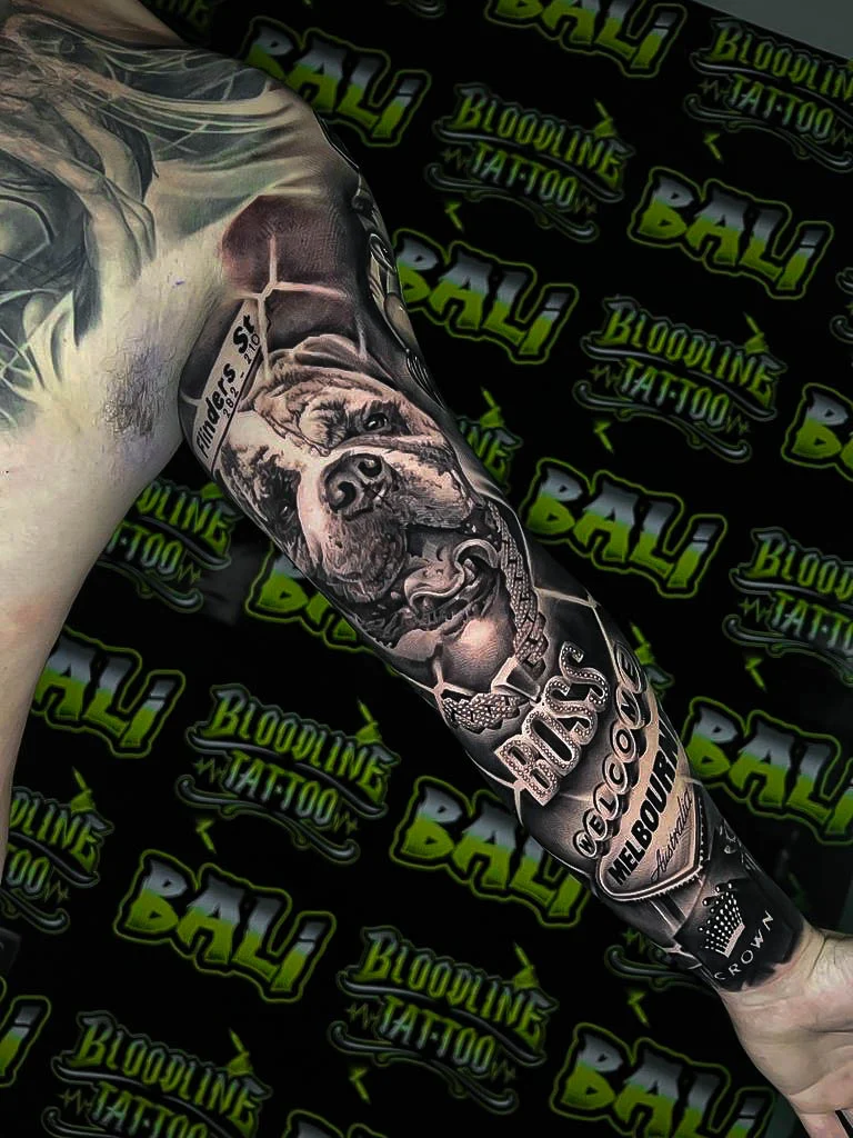 Black and grey portrait realism arm tattoo of a bulldog memorial portrait with script, jeweled chain, and bottle label details at Bloodline Tattoo Bali.