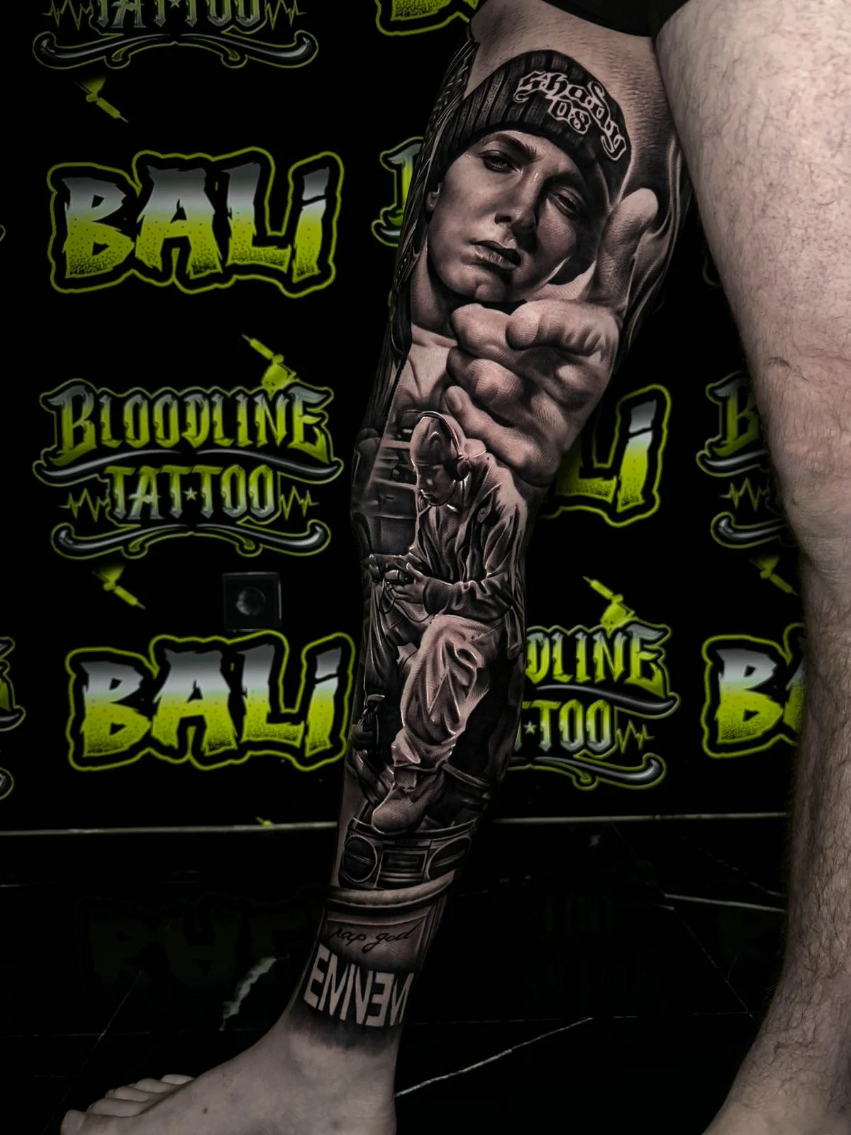 Black and grey portrait realism leg tattoo of Eminem with a beanie, pointing hand, seated performance scene, and name lettering at Bloodline Tattoo Bali.
