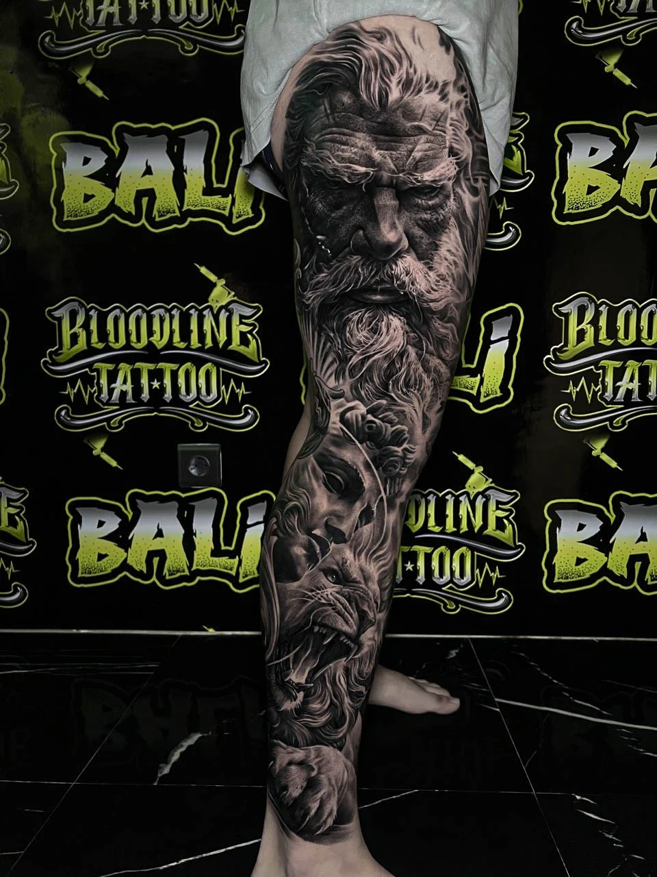 A black and grey realism lower leg sleeve of a solemn bearded face emerging through layered leaves and smoky organic textures, at Bloodline Tattoo Bali.