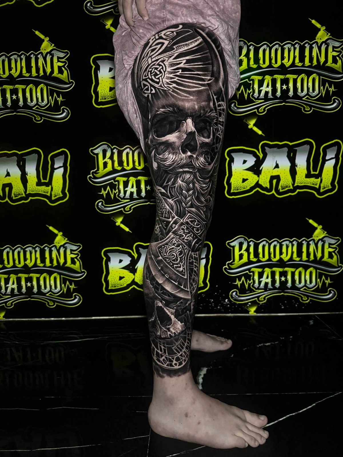 A black and grey realism lower leg sleeve featuring a stern warrior portrait with ornate filigree and textured darkwash shading, at Bloodline Tattoo Bali.