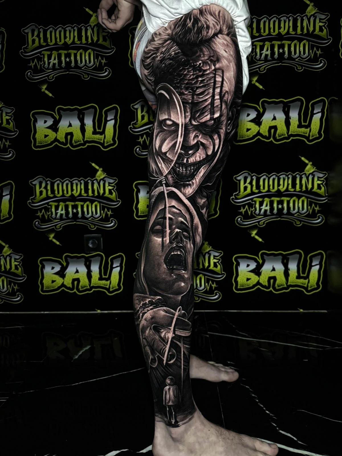 A black and grey realism lower leg sleeve with a sinister grinning clown, a shadowed female face, and smoky horror shading, at Bloodline Tattoo Bali.