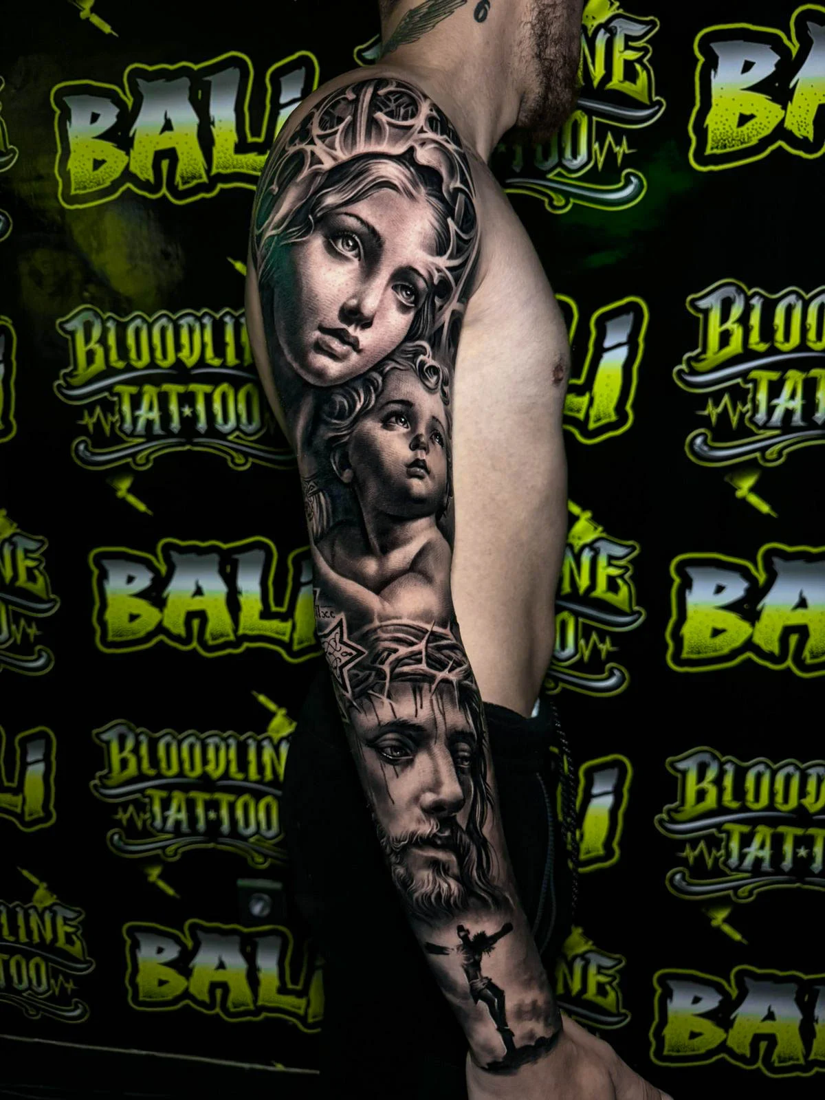 A black and grey realism arm sleeve with a sorrowful woman's face, a child's portrait, and a dark sacred face layered beneath, at Bloodline Tattoo Bali.