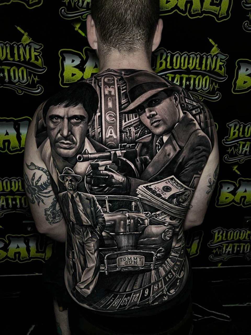 A black and grey realism full back piece with Scarface, a gun-wielding woman, cash, and gangster props rendered in sharp noir contrast, at Bloodline Tattoo Bali.