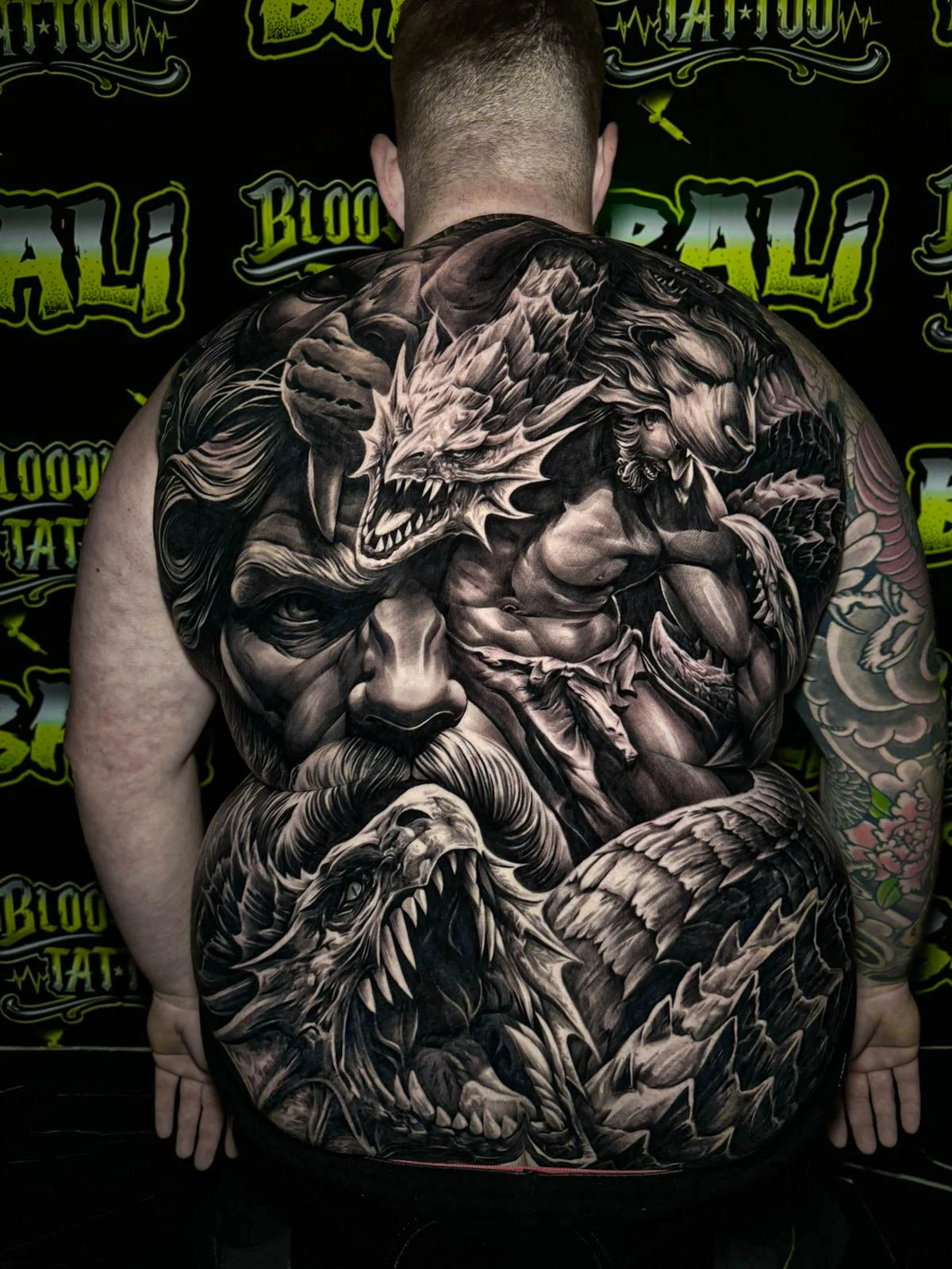 A black and grey realism full back piece with a commanding bearded king, a dragon, and layered eagle feathers in powerful dark contrast, at Bloodline Tattoo Bali.