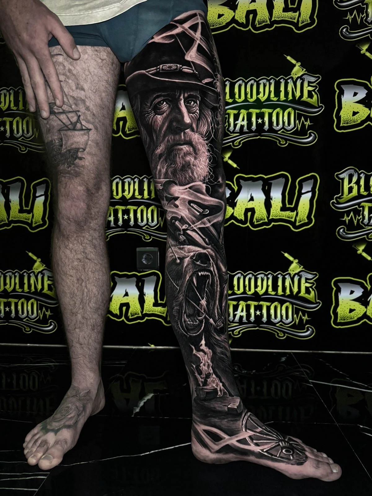 A black and grey realism lower leg sleeve of an elderly bearded portrait with weathered skin texture and moody darkwash shading, at Bloodline Tattoo Bali.