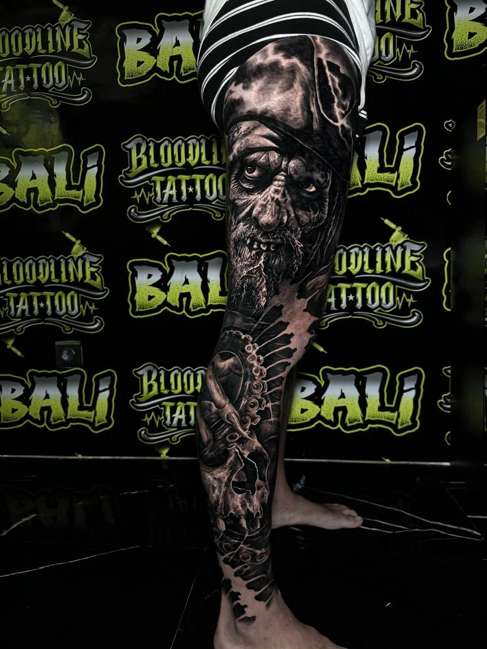 A black and grey realism lower leg sleeve with an elderly bearded face, feminine portrait detail, and soft smoke transitions, at Bloodline Tattoo Bali.